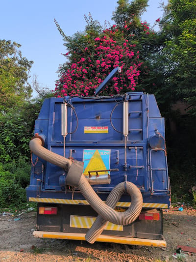 The image shows the rear of a blue garbage truck parked outdoors. The truck has a large, corrugated hose attached to its left side, extending downwards and looping outwards. There are several stickers on the back of the truck, including one that reads "PYARA SINGH & SONS" and another with the words "SLOW" in large yellow letters. The truck is surrounded by lush green foliage and a tree with bright pink flowers. The ground is a mixture of dirt, gravel, and some scattered debris, suggesting an unkempt environment. The sky above is clear blue, indicating it is daytime. The lighting suggests it is late afternoon, with long shadows cast on the ground. The scene appears to be in an outdoor area, possibly a junkyard or a less maintained part of the city, amidst natural vegetation. There are no people visible in the image. The presence of the "PYARA SINGH & SONS" sticker strongly suggests the location is in India, and given the context of previous images, it is likely in the city of Jammu, India.