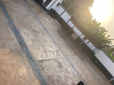 The scene, located in Ogbunu, Nigeria, depicts an outdoor area under direct sunlight, with a pronounced diagonal tilt. The foreground features an expansive, patterned concrete surface primarily in shades of light brown and tan. This surface is segmented by dark grey strips, creating distinct rectangular and square sections. These sections exhibit various embossed textures, including simulated wood grain, geometric patterns, and pebble-like designs. To the right, a low wall or fence with a white upper section and a dark grey base runs parallel to the patterned ground. Above this wall, a dark, potentially corrugated metal or thatched awning extends over a portion of the structure. Behind this wall and awning, dense green tropical foliage, identifiable as large banana leaves, is visible. In the background, further structures or buildings with white and dark sections are partially discernible. The sun, positioned in the upper right, casts significant glare and bright illumination across the scene. A black cylindrical object is partially visible in the lower left corner. No human figures are present.