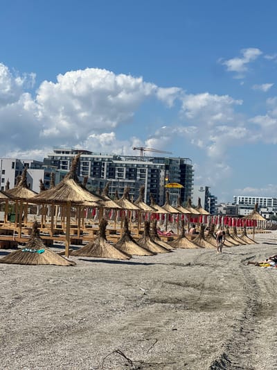 The image shows a beach in Năvodari, Romania, with a row of thatched umbrellas and lounge chairs. In the background, modern apartment buildings line the coast, with a construction crane visible among them. A few people are scattered on the beach, with one person standing near the umbrellas and another lying down on a lounge chair. The sky is blue with scattered white clouds. The beach appears to be made of sand and small pebbles.