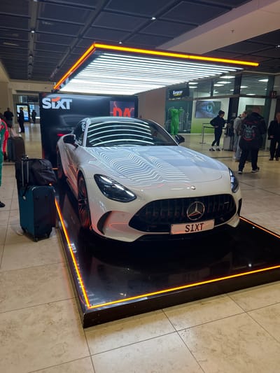 The image presents an indoor scene, likely an airport terminal or a large commercial area, based on the prominent car rental branding and visible luggage. The central subject is a white Mercedes-Benz sports car, possibly an AMG GT, showcased on a raised, rectangular black platform. This platform is strikingly outlined with bright orange-red LED light strips, drawing attention to the vehicle. The car itself features a prominent Mercedes-Benz star logo on its grille and a front license plate clearly displaying "S1XT."

Behind the car, a tall black banner or wall features the orange "SIXT" logo, indicating the car rental company responsible for the display. In the background, further signs are visible, including a "Europcar" logo with yellow text on a green background. There is also a large screen or poster showing a close-up of an eye in blue tones, and another green graphic with a stylized cityscape and a frog-like figure.

Several people are scattered throughout the background, appearing to be travelers or visitors observing the display or moving through the space. Some luggage, including a black backpack and a blue wheeled suitcase, rests on the light-colored tiled floor near the car. The environment is brightly lit by numerous long, linear ceiling lights, whose reflections are visible on the car's sleek surface. The scene suggests a promotional display within a modern, well-lit public space, consistent with an airport setting in Schönefeld, Germany. No specific emotions are evident, and the time of day is not distinguishable due to the artificial lighting.