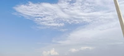 The image displays a bright, overcast sky with a gentle gradient from a clear blue at the top to a hazy, lighter tone towards the horizon. Wispy clouds are scattered across the upper portion of the frame, with denser formations of white and grey clouds dominating the middle and lower sections. A tall, slender, light-colored pole, possibly a utility pole or a flagpole, is visible on the right side of the image, extending vertically from the bottom edge towards the top. There are no people or distinct objects on the ground, suggesting an elevated perspective or a focus solely on the sky. The overall impression is one of a calm, perhaps slightly humid, atmospheric condition.