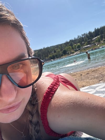 A woman lies on a beach in the city of Maisod, France, by a lake. She is wearing sunglasses and a red bikini. Her hair is braided. The water is a clear blue, and there are trees and buildings in the background. The sky is a bright blue.