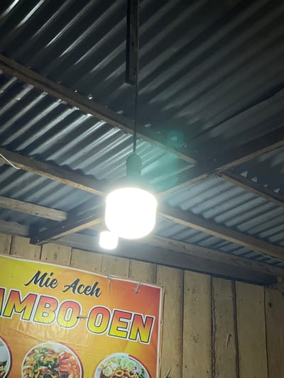 This image depicts an interior scene in Alue Mudem, Indonesia, characterized by a utilitarian structure. The upper portion of the frame shows a grey, corrugated metal roof supported by rough-hewn wooden beams. From a central wooden support, two bright white LED light fixtures are suspended by dark wiring: a larger, cylindrical main light and a smaller, auxiliary light positioned below it. The intense illumination from these fixtures creates significant glare and a faint greenish lens flare. The right and lower portions of the image reveal a wall constructed from vertical wooden planks, exhibiting an aged, light brown appearance. Affixed to this wooden wall is a partially visible banner, yellow and orange in color, advertising "Mie Aceh JUMBO-OEN" with depicted images of noodle dishes. Multiple small geckos, approximately five to six, are observed adhering to both the wooden wall and the surface of the banner. No human subjects are present.