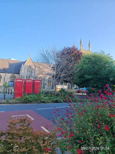 The image depicts a street scene in Eastbourne, United Kingdom. In the foreground, a patch of red flowers and green foliage lines the edge of a road. The road itself is asphalt with a painted white line. To the left of the road, a maroon-colored paved area with yellow arrows is visible.
Further back, three iconic red telephone boxes stand in a row, set against a backdrop of lush green bushes and trees. Behind the telephone boxes, a stone building with gothic architectural features, likely a church, is prominent. The building has a dark, sloping roof and arched windows. Two spires, characteristic of a church tower, rise above the roofline against a clear blue sky. A bare-branched tree stands to the right of the building.
The overall scene is bathed in the warm light of what appears to be either early morning or late afternoon. A timestamp "08/07/2026 19:34" is overlaid in the bottom right corner of the image. No people are visible in the frame.