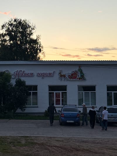 The image shows a building facade in the city of Kukui, Russia, decorated with a "Happy New Year!" inscription in Russian Cyrillic, alongside a Christmas-themed decal featuring Santa Claus in a sleigh pulled by a reindeer, and a decorated Christmas tree. The building appears to be an institutional or commercial structure, with several windows visible. The sky at dusk displays soft pink and orange hues. In front of the building, a group of four men are standing near parked cars. One man, in the foreground to the left, is walking away from the building. Two other men are standing in the middle, near a blue car, and another man is standing to the right, near a silver car. The ground in front of the building is paved, with some grassy areas visible. The overall scene suggests a late evening in a town or city.