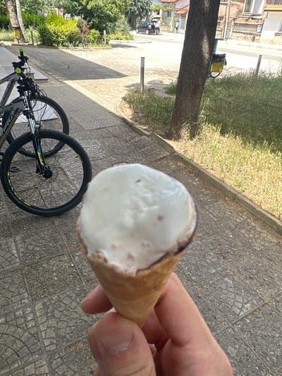 The image shows a close-up of a person's hand holding a partially eaten ice cream cone. The ice cream appears to be vanilla flavored with small brown speckles, possibly chocolate chips or nuts. The cone itself is light brown and appears to be a waffle cone. The background is blurred but shows a paved walkway with square tiles, a tree with rough bark, and some greenery, including grass and bushes. In the lower left portion of the image, a bicycle is visible, with its front wheel and handlebars in focus. The overall scene suggests an outdoor setting, likely on a sunny day, given the bright lighting and shadows. The presence of the bicycle and the casual nature of the ice cream cone suggest a leisurely activity, possibly a break during a bike ride or a walk. The location is identified as Ovoshtnik, Bulgaria. There is no visible text in the image.