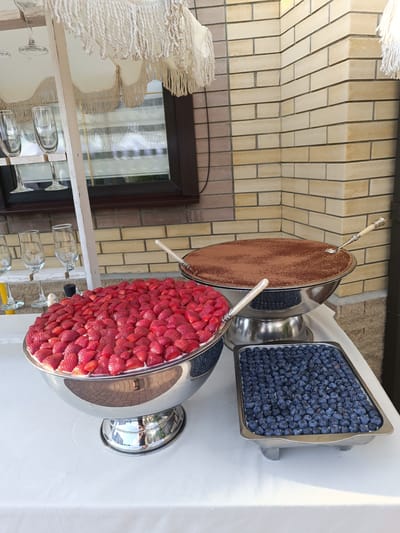 In Pchegatlukay, Russia, an outdoor table is set with various desserts, presumably for a gathering. The table is covered with a white tablecloth, upon which rests a large silver bowl filled to the brim with fresh, bright red strawberries. To the right of the strawberries, a rectangular metal tray is packed with blueberries. Behind the strawberries, a very large, shallow silver bowl contains a dessert dusted with cocoa powder, possibly tiramisu. Two ornate serving spoons, one with a light-colored handle and another with a darker handle, are placed in the desserts. In the background, a light-colored brick wall forms the backdrop. To the left, a white wooden structure, resembling a shelf or ladder, displays several empty stemmed glasses. The scene appears to be set up for a celebration or event, with the natural light suggesting it is daytime.