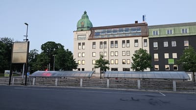 The image shows the exterior of a building complex located in Jönköping, Sweden, specifically near the central train station and the nearby bus terminal. The main building features a white facade with multiple stories of windows, topped with a green dome and a glass-enclosed upper level. Adjacent to it is a dark grey building with a green roof, also featuring multiple windows, and a visible "DIVID" logo. In front of the buildings, a bus shelter with a transparent roof is situated over what appears to be bicycle parking. The foreground consists of an asphalt road. A public information display board, showing schedules, is visible to the left, along with trees and streetlights. The sky is a pale blue, indicating it is either early morning or late evening. There are no people visible in the image.