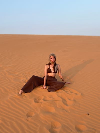 A single female subject is seated centrally on reddish-orange sand dunes, consistent with the landscape of the Red Sand Dunes in Phan Thiet, Vietnam. She is positioned with her legs splayed, the left leg extended forward and the right leg bent at the knee, both bare feet partially visible in the sand. Her body is angled slightly to the left, and she faces the viewer directly with a neutral expression. She is dressed in a dark brown, two-piece ensemble consisting of a bikini-style top and loose, wide-leg pants. A light beige or cream-colored headscarf is worn, covering her head and tied under her chin. A dark, serpentine tattoo is visible on her left forearm. Her hands rest on the sand beside her. The surrounding environment comprises extensive, undulating sand dunes with prominent wind-formed ridges and troughs. A clear, light blue sky meets the sand at a sharp, level horizon line. An elongated shadow of the subject is cast to her right on the sand, indicating a low sun angle. Several footprints are discernible in the sand in the lower left foreground.