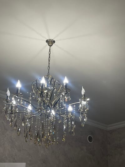 A low-angle shot focuses on a lit crystal chandelier. The chandelier is suspended from a chain attached to a ceiling mount. The ceiling is white and marked with shadow. The walls are gray. A circular recess light is mounted. A wall edge is visible. The location cannot be determined.