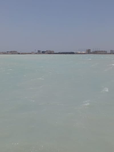 The image depicts a view of the waterfront in Al Khobar, Saudi Arabia, from the sea. The buildings of the city are visible in the distance, blurred due to the distance and atmospheric conditions. The foreground is dominated by the water, which is a pale blue-grey color, showing gentle ripples. The sky is clear and pale blue. No people or other objects are clearly visible in the scene.