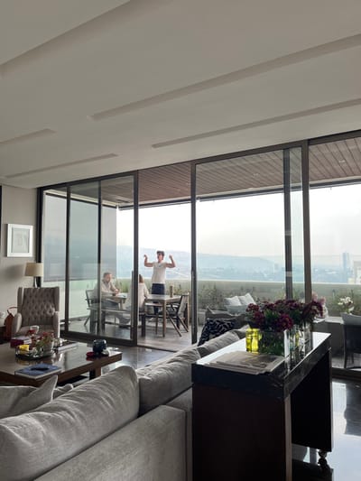 An elevated view from a modern apartment in Santiago Yancuitlalpan, Mexico, shows an interior living space opening onto an outdoor balcony. In the foreground, a light gray sectional sofa and a dark wood coffee table with a silver tray, red flowers, and a blue decorative object are visible. To the left, a gray upholstered armchair and a floor lamp stand near a white-framed picture on a light wall. Behind the sofa, a dark marble-topped console table holds magazines and vases with yellow liquid and purple flowers.
Through expansive glass sliding doors, the outdoor balcony is visible, furnished with a dark wooden dining table and chairs, and a dark outdoor sofa with pillows. Three individuals are on the balcony. A young male in a white t-shirt and dark shorts stands centrally, facing the camera, flexing both biceps. To his left, a bearded male and a female with lighter hair are seated at the dining table, engaged with objects on its surface, suggesting a game or activity. The balcony floor is light wood decking, and potted plants line the railing. Beyond the balcony, a hazy cityscape with buildings and distant green hills/mountains extends under a bright sky. The interior ceiling is white with linear recessed panels.