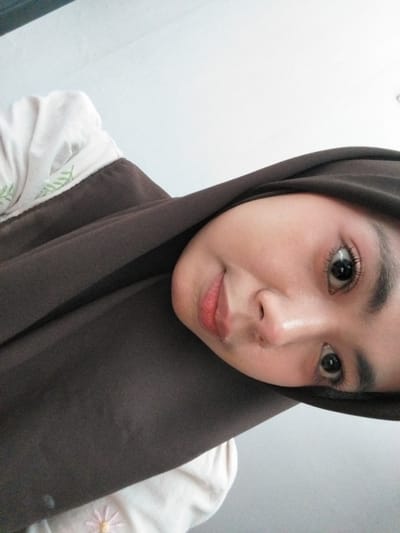 A close-up, vertically oriented selfie shows a young woman wearing a dark brown hijab and a light-colored shirt with green embroidered leaves. Her face is tilted towards the camera, with her left eye more prominently displayed. She has dark eyebrows, long eyelashes, and light pink lips. The background appears to be a white wall. The image was taken in Tibojong, Indonesia.
