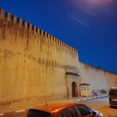 The image depicts a street scene in Mechouar Fes Jdid, Morocco, at night. A high, ochre-colored wall with crenellations runs along the background. A doorway with a green-tiled roof juts out from the wall. To the right of the doorway is a small, yellow, booth-like structure. Two people stand next to a stroller, while a third person sits on a white chair. An orange car is parked on the left side of the street, and a dark-colored van is parked on the right. The sky is a deep blue.
