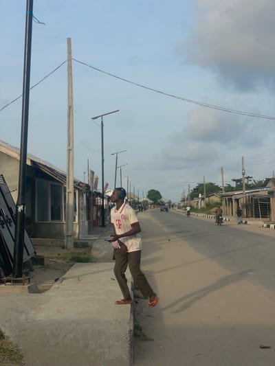This image depicts a street scene in the city of Mosafejaw, Nigeria, under an overcast sky. In the foreground, a young man is positioned on the edge of a concrete sidewalk, facing left and looking upward. He wears a white polo shirt with a red collar and "T" logo, dark trousers, and orange flip-flops. Black over-ear headphones are on his head, and he holds a smartphone in his left hand. His body is angled, and his left foot is on the sidewalk while his right foot is suspended just above the dirt shoulder, casting a long shadow onto the street.
To the man's left, a black metal pole with a blue cord attached rises in the immediate foreground, alongside a lighter, bare utility pole. Behind these, a row of single-story buildings or stalls with varied facades, including corrugated metal and glass windows, line the left side of the street. Multiple black streetlights, featuring solar panels, are visible along this side, extending into the distance.
The paved road, appearing to be asphalt or tar, stretches into the background, flanked by dirt shoulders. On the right side of the road, more buildings of varying construction materials are present, along with several utility poles supporting power lines. Green foliage is visible further down the street.
In the mid-ground, a light-colored car is seen driving away from the viewer, and two individuals on motorcycles are also observed heading in the same direction. A solitary figure walks on the right shoulder further down the road. The overall environment suggests a semi-urban or suburban area with ongoing development and daily activity.