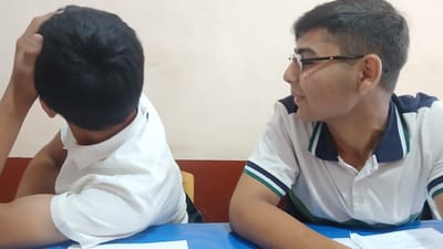 Two young males are seated at a blue table in what appears to be a classroom or study hall. The male on the left is viewed from the back, with his left arm raised and hand on his head. He is wearing a white collared shirt. The male on the right is facing to his left, looking towards the other male. He is wearing glasses and a white collared shirt with dark blue and green trim. The background consists of a light-colored wall with a red stripe near the bottom. The scene is likely set in Kattakurgan City, Uzbekistan.