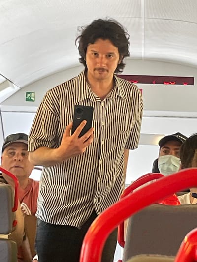 The image depicts a scene inside what appears to be a train or bus, possibly in or near the city of Garraf, Spain, given the presence of individuals who may be locals or tourists. A man with dark, curly hair and a mustache is holding up a smartphone, likely taking a picture or video. He is wearing a striped shirt and black jeans. In the background, another individual wearing a black baseball cap and a face mask is visible. Another person is visible in the bottom portion of the image in a red shirt and black hat. Red bars of the seats are visible. The interior has white walls and a red electronic signage panel. A green exit sign is attached to the wall. The camera is capturing this moment from the perspective of someone either sitting near or involved in the scene.