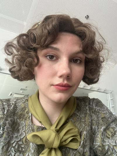 A light-skinned female individual is depicted in a head-and-shoulders portrait, facing directly towards the camera. She possesses shoulder-length, dark brown hair styled in tight curls, brown eyes, and wears light pink lipstick. Her attire includes a top or dress featuring a muted gold or olive-green base overlaid with greyish-brown lace, and an olive-green satin scarf tied in a prominent bow around her neck. The background is out of focus, revealing an indoor environment with a white ceiling composed of perforated acoustic panels. To the right, a mirror framed with multiple spherical light bulbs is partially visible. A white rectangular vent is present on the left side of the background. The precise location within Huangdao District, China, cannot be determined from the visual information provided.