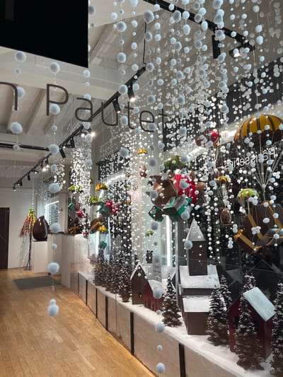 A retail display features hanging decorations: spheres resembling snow and various chocolate-themed objects like hot air balloons, gift boxes, and houses. The display is visible through a glass window, reflecting the interior of the store, including a wooden floor. The name "pâtisserie" is partially visible.