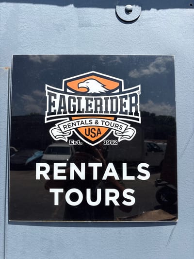 The image shows a black sign with white and orange lettering affixed to a light blue, textured wall. The sign displays the "EAGLERIDER" logo, which includes an eagle's head and the words "RENTALS & TOURS" and "USA". Below this, it says "Est. 1992". The bottom portion of the sign reads "RENTALS TOURS" in large white capital letters. Reflections on the sign indicate the presence of vehicles and possibly people. The sign is mounted on an exterior wall of a building, possibly a garage or service area. The overall scene suggests a commercial establishment. The location is the EagleRider motorcycle rental and tour company, located at 1340 Rand Road in Des Plaines, Illinois, United States of America.