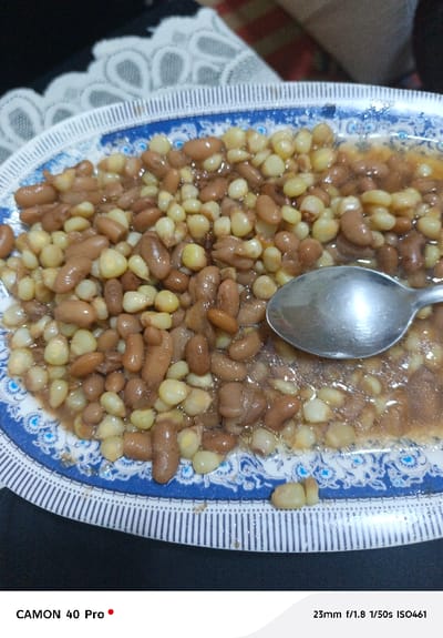 The image displays a close-up, overhead view of a meal consisting of githeri (a mixture of maize and beans) served in a round ceramic plate. The plate features a distinctive blue and white pattern, with a striped rim and an inner band of blue floral or swirling motifs on a white background. A silver-colored metal spoon rests within the food, partially submerged in the liquid sauce or broth accompanying the maize and beans. The food itself is a homogeneous mix of light brown, kidney-shaped beans and pale yellow corn kernels. The plate is positioned on a dark, possibly black, surface, likely a table. A white, intricately patterned lace tablecloth or mat is partially visible in the upper left background, and a blurred reddish-brown texture is present in the upper right. The scene is located in Kitengela ward, Kenya. No individuals are visible, and no active interactions or actions are depicted.