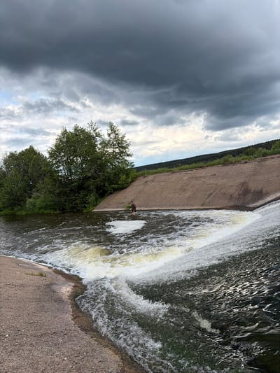 The image shows a waterfall or dam in Nizhnii Vishur, Russia. Water cascades down a concrete structure into a pool below. There's a person standing in the pool near the base of the waterfall. Lush green trees are visible along the left side of the concrete structure. The sky is dominated by ominous dark storm clouds.