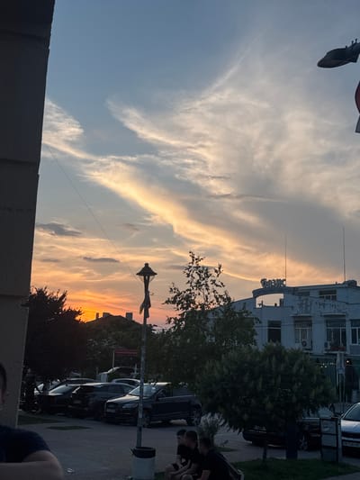 The image captures a sunset scene in the city of Rakovski, Bulgaria, characterized by an orange and red horizon transitioning to a light blue sky with elongated clouds. In the immediate lower-left foreground, the shoulder and arm of a person, clad in a dark shirt, are partially visible. A dark grey building facade occupies the left edge of the frame. In the lower-midground, three young men are seated on a low structure, wearing dark attire and observing the scene to their right. A black street light pole with a decorative fixture and a small banner stands centrally, accompanied by a dark trash receptacle at its base. A paved area containing multiple parked cars, including a dark SUV with a visible partial Bulgarian license plate (e.g., PB 3448XX) and a white car with plate PB 7580 PE, extends into the mid-ground. Dense green foliage and trees are interspersed throughout the mid-ground. In the background, a light-colored, two-story building with peeling paint or wear features a prominent Cyrillic sign reading "БАЗИС" (BASIS) on its roof, from which two thin antennas extend. The sun is visibly setting behind distant structures and trees on the left side of the frame.