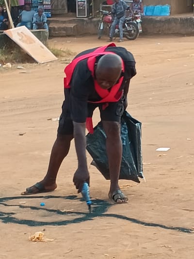 In an outdoor shot, a person wearing a black shirt, shorts, sandals, and a red safety vest is bent over drawing a design on the sandy ground with a blue tool. A black bag hangs near the person's legs. In the background, people are seated, and a motorcycle is parked nearby, further away in the distance, is a building. The lighting suggests it could be daytime.