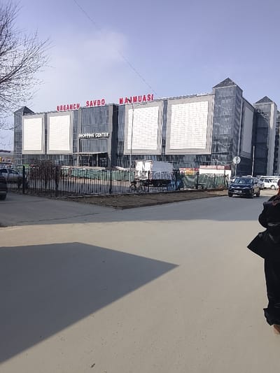 The image depicts a daytime scene in Urgench, Uzbekistan, focusing on a modern shopping center. The prominent building, identified by the red signage "URGENCH SAVDO MAJMUASI" and a central "SHOPPING CENTER" sign, features a facade of dark grey panels and extensive glass, including two pyramidal glass structures at its upper corners. Large, light-colored rectangular panels are integrated into the building's design.
In the foreground, a paved road or large sidewalk occupies the lower half of the frame. A low, black metal fence separates this paved area from a patch of barren earth and the shopping center's immediate vicinity. Behind the fence, a white cargo van or truck is parked alongside a bicycle. Several other vehicles, including a dark-colored SUV, are visible on the road to the right, with additional parked cars further in the distance. A bare tree branch extends into the upper left of the frame. A partial figure, clad in dark clothing, is visible on the far right edge of the image. Sunlight casts distinct shadows on the paved ground. The sky is clear and light blue.