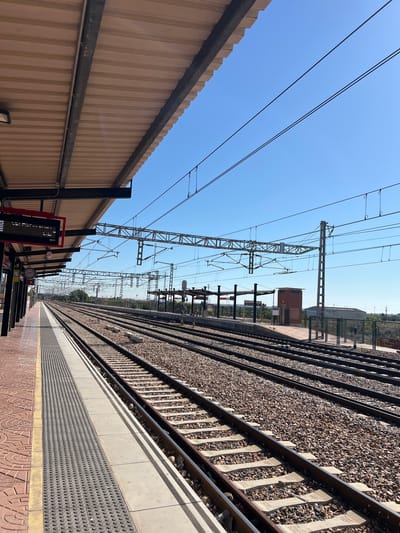 The image captures a railway station in Puçol, Spain. The viewpoint is from the platform, looking lengthwise along the tracks. On the left is a corrugated metal fence, and overhead are electrical wires. To the right, parallel train tracks run along gravel, with a gray safety strip with raised dots along the platform edge. The platform surface is patterned with red and brown tiles. In the distance are buildings and greenery. The sky is clear and blue. The perspective creates a sense of depth, emphasizing the station's length.