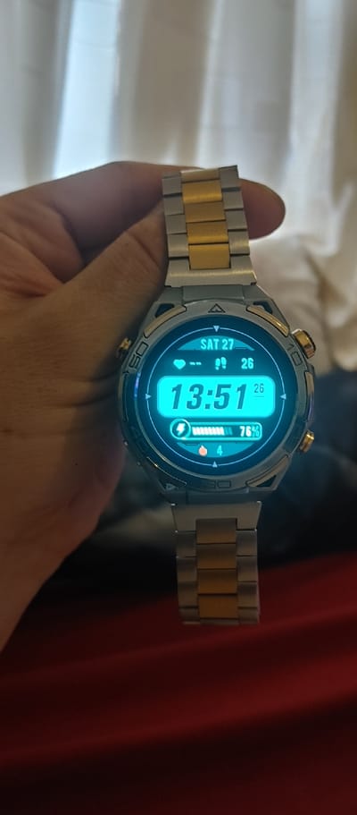 A person's hand is holding a smartwatch with a metal band composed of alternating gold and silver links. The watch face is a bright blue and displays digital information. The time reads "13:51". Above the time, "SAT 27" is visible, indicating the day and date. To the right of the time, a battery icon shows "76%" remaining. There are also small icons that appear to be for steps and heart rate. The watch has a circular bezel with markings, including "OS" and "30" visible on the side. The background is blurred, with hints of white fabric and red drapery. The scene appears to be indoors, possibly in a home environment, given the fabrics. No specific weather or time of day can be determined beyond the watch display. The overall impression is a close-up shot of a modern piece of technology being displayed.