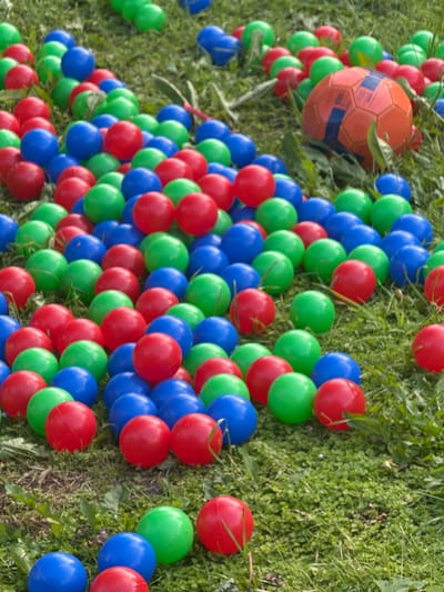 A collection of plastic balls and one soccer ball are scattered on a grassy lawn. The balls are primarily red, green, and blue, with one orange soccer ball featuring blue accents. The image appears to be taken outdoors in daylight, with shadows indicating the sun is present. The ground is covered in green grass and some scattered weeds. The context suggests a recreational or play area, possibly a backyard or park, in Дзержинец-83 Снт, Russia. There are no people visible in the image.