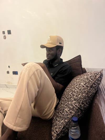 A young man wearing a tan baseball cap and a black t-shirt is lying on a brown couch. He is wearing light-colored sweatpants. A patterned pillow is behind him, and a plastic water bottle with a blue cap is on the couch next to the pillow. The room has a white wall with electrical outlets. The lighting suggests it is daytime. The man's expression is relaxed. The text on his cap reads "EIN".