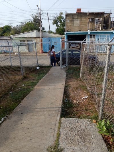 The image shows a street scene in Barcelona, Venezuela. A concrete sidewalk leads into a gated area with a chain-link fence on either side. A woman wearing a white cropped top, jeans, and a black cap is standing near the gate. A dark-colored car is visible through the gate. Behind the gate is a building painted blue with a second-story addition constructed with concrete blocks. A smaller, white building is visible in the background to the left. Overhead power lines are present, and there is scattered litter on the ground. The area appears unkempt.