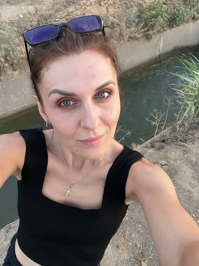 A woman with blue eyes and brown hair pulled back is taking a selfie outdoors. She is wearing a black tank top and a silver necklace with a cross pendant. Sunglasses are perched on top of her head. She is standing on a dirt path next to a canal with green water. The background shows dry vegetation and a concrete structure. The location is the city administration area of Aqsu city, Kazakhstan.