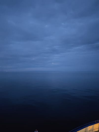 The image depicts a dark, expansive body of water under a cloudy, twilight sky. A thin sliver of a crescent moon is visible in the upper portion of the frame. In the distance, a faint line of lights suggests a shoreline or a settlement. The foreground shows the edge of a boat, with a blue railing and yellow lighting. A small, indistinct figure of a person is visible near the bottom center, standing on the boat. The overall impression is one of a serene, possibly cold, maritime environment at dusk. The image is attributed to Shcheleiki, Russia, suggesting a location on a coast or large body of water within that region.