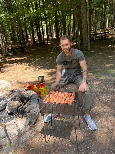 The image depicts an outdoor scene in a sunlit forest clearing in Mali Bostur, Montenegro. An adult male is seated on a wooden log, facing the camera with a smile, while grilling meat skewers on a rectangular metal grill. He wears a dark olive green t-shirt with "STAY UP" printed in capital letters, matching olive green cargo pants, and light grey Adidas athletic shoes with red accents. A multi-colored tattoo covers his left forearm. Approximately ten skewers with raw meat and onion pieces are visible on the grill, positioned over coals. To the immediate left of the grill, a stone-lined fire pit contains charred wood and ash. Behind the fire pit are two yellow and red patterned bags, one partially open, and a silver pot with a lid resting on the ground. A green glass bottle is partially visible in the bottom left foreground. The ground consists of dry earth, scattered fallen leaves, and small rocks, with dappled sunlight and shadows cast by numerous tall trees with green foliage. In the background, two wooden picnic tables with benches are visible under the tree canopy.