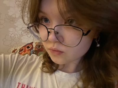 A person with blue eyes and glasses is pictured close-up. They wear a white t-shirt with red letters and a necklace. The background shows a floral-patterned surface and a colorful pillow with an owl design. One earring is visible on the left side of the image. The picture is rotated.