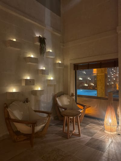 A dimly lit interior space in Ürgüp, Turkey is shown. The space features stone walls with embedded rectangular lights. A cluster of herbs is fixed on one of the stones. Two wooden rocking chairs with white cushions sit near a window that looks out onto a pool area illuminated with lights. Above the chairs hangs a wicker lampshade. The floor is tiled in a diamond pattern.