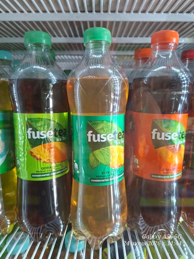 The image shows a close-up of three bottles of Fuse Tea ice tea in a refrigerator. The bottles are arranged in a row, with their labels facing forward. The left bottle has a green cap and a label with an image of a pineapple and green leaves. The middle bottle also has a green cap and a label with a similar design, featuring pineapple and leaves. The right bottle has an orange cap and a label with an image of a peach and green leaves. The liquid inside the bottles appears to be a dark amber or brown color. The bottles are placed on a white slatted shelf. In the bottom right corner of the image, text reads "Galaxy A56 5G 24 июня 2026 г. 11:44". The setting is indoors, likely a commercial refrigerator in a store or market. No people or specific urban landmarks are visible.
