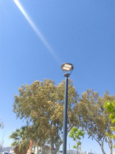 The image depicts a clear blue sky with a bright sun flare streaking across the upper left portion. In the mid-ground, a dark gray, modern lamppost with a circular head containing two rectangular light panels stands prominently. Behind the lamppost, a dense canopy of deciduous trees with light green and yellow foliage fills the lower half of the frame. To the left of the lamppost, a portion of a palm tree with green fronds is visible. In the far background, faint outlines of buildings and hills can be discerned. The overall scene suggests an outdoor public space, possibly a park or a street, in the Municipal Unit of Elefsina, Greece.