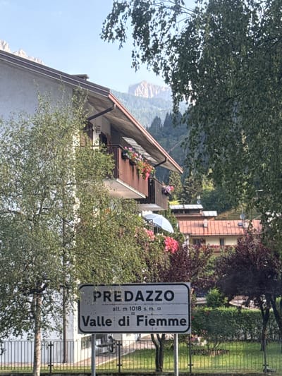 The image shows a sign for Predazzo, Valle di Fiemme, at an altitude of 1018 meters. The sign is in the foreground, with a fence and a grassy area behind it. In the background, there are buildings, trees, and mountains. The sky is blue with some clouds. The scene appears to be a roadside view in a mountainous region of Italy.