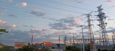 This is an exterior, eye-level, medium shot of a commercial and industrial area in Sagunto, Spain, at dusk. The sky is filled with scattered clouds, tinged pink by the setting sun, against a pale blue backdrop. Several high-voltage electricity towers dominate the right side of the frame, their lattice structures silhouetted against the sky. Power lines stretch across the upper portion of the image.
In the foreground and midground, there are commercial buildings. A cylindrical building with the illuminated red logo "YELMO CINES" is visible on the left. Next to it, a rectangular building features a large illuminated sign that reads "Kyoka RESTAURANTE" with a red circular logo. The buildings have modern facades with horizontal bands of color.
To the left of the frame, trees and foliage obscure the lower part of the commercial structures. Streetlights with red poles and dual lamps are positioned in front of the buildings. In the background, a row of smaller electricity pylons and substations can be seen. The overall scene depicts a blend of urban infrastructure and commercial development under a dramatic evening sky.