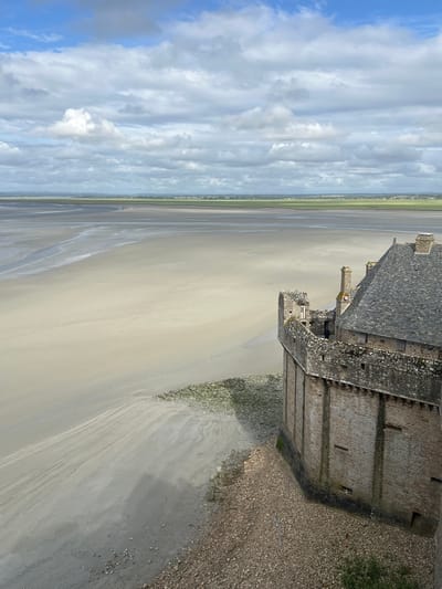 A high-angle, eye-level shot captures a view from the ramparts of Mont Saint-Michel, France, looking out over the vast expanse of the bay. The foreground is dominated by the textured stone wall and weathered grey slate roof of a medieval structure, likely part of the abbey or its associated buildings. The wall is constructed from rough-hewn stone blocks, with darker, moss-covered sections indicating age and exposure to the elements. Narrow windows and small openings are visible along the wall's surface. Beyond the wall, a wide, flat, sandy foreshore stretches into the distance, marked by subtle ripples and streaks suggesting tidal movement. In the far distance, a thin line of green vegetation marks the mainland coastline, under a sky filled with dramatic, puffy white clouds against a pale blue background. The overall scene conveys a sense of historical grandeur and the raw beauty of the natural environment at low tide.