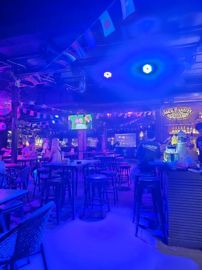 This is a medium shot, taken from a slightly low angle, of the interior of a bar in Ban Klong Haeng, Thailand, illuminated primarily by blue and purple lighting. The scene is populated by several patrons, mostly seated at tables or standing near the bar. The bar itself is well-stocked with bottles of alcohol, including prominently displayed Jack Daniel's signage. A television screen in the background shows a sports broadcast. The ceiling is adorned with numerous small flags, suggesting an international theme or a celebration of a sporting event. The overall ambiance appears lively and casual, typical of a bar or pub environment.