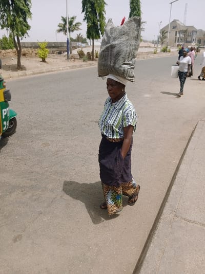 The image portrays an outdoor street scene in Kadoma, Zimbabwe, captured on a bright, clear day. In the foreground, slightly left of center, an adult female with dark skin walks forward, angled slightly to her right. She wears a white head covering, a short-sleeved collared shirt with horizontal white and light green stripes, a dark blue or black skirt with a patterned lower hem, and dark flip-flops. A large, grey, woven sack, containing a red item at its peak, is balanced on her head. Her right hand is obscured, possibly in a pocket. To her immediate left, the front portion of a green and yellow auto-rickshaw, displaying "PT" and "39," is partially visible. The paved road surface extends into the background, bordered on the right by a raised curb. Further down the road, another adult female, dressed in a white t-shirt and patterned leggings, carries a white bucket. Other indistinct figures are visible in the distant background. The environment includes several tall, slender green trees, a palm tree, and various bushes lining the road. A concrete bridge structure is present in the mid-ground. In the far background, multiple buildings, including a multi-story structure, and utility poles are visible. Shadows are cast to the right of the main subject, indicating a high sun position.