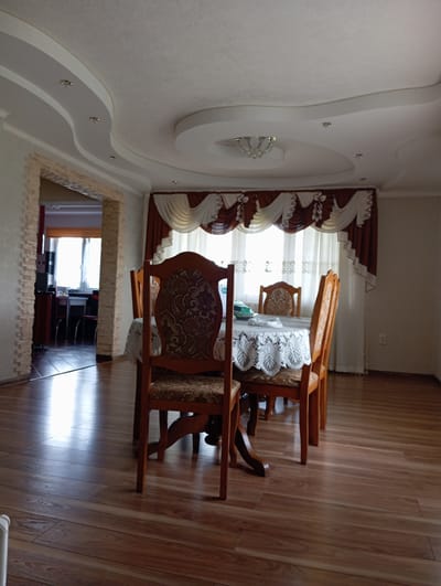 The image depicts an interior scene, likely a dining room, within a residential property located in Hlynytsi, Ukraine. The room features a dining table covered with a white lace tablecloth, surrounded by four wooden chairs with patterned upholstery. The floor is made of polished wood laminate. A large window with layered curtains, featuring a brown and white decorative trim, is visible behind the table. The ceiling is adorned with an intricate plaster design, incorporating curved elements and recessed lighting fixtures, as well as a central chandelier. An archway on the left side of the image leads to another room, revealing a glimpse of a window and furniture. The overall ambiance is domestic and appears to be well-maintained.
