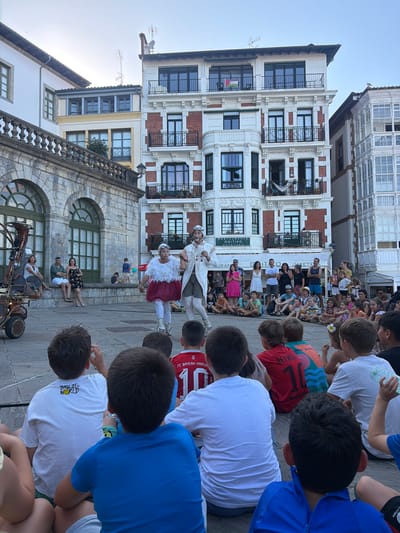 In Lekeitio, Spain, a street performance is underway in a public square with a stone building on the left and ornate apartment buildings in the background. The focus is on two performers dressed in elaborate costumes, one in white with a feathered collar and the other in a white coat, white leggings, and goggles. They are performing for a crowd of onlookers, primarily children seated on the ground in the foreground, facing away from the camera. The children are wearing casual clothing, with some wearing soccer jerseys. Other spectators, adults and children, are visible further back, some standing and some seated on a stone ledge. A large, steampunk-like mechanical structure is partially visible on the far left. The atmosphere appears festive, with the performance drawing attention from the gathered audience.