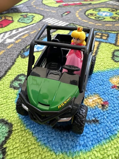 A toy John Deere Gator RSX860i utility terrain vehicle (UTV), predominantly black with a green hood and yellow "RSX860i" lettering, is positioned on a multicolored play rug. A plastic figure, identifiable as Princess Peach from the Super Mario Bros. franchise, wearing a pink dress and a gold crown, is seated in the driver's seat of the toy UTV. The rug features a road map pattern with gray roads, green land, and blue water elements. A blurry, light-skinned limb is partially visible in the upper left corner of the frame. The scene is indoors. The precise location within Naperville, United States, cannot be determined from the visual information.