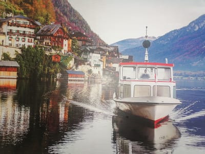 The image depicts a serene waterfront scene, featuring a white passenger boat with a red trim navigating through calm water. The boat, positioned in the foreground and to the right of the frame, is creating gentle ripples. Behind the boat, a picturesque village is nestled on a steep hillside, characterized by traditional wooden houses with red roofs, interspersed with autumnal foliage. The village buildings have multiple stories and balconies, with some appearing to be historically preserved. The hillside is covered in trees displaying vibrant fall colors, ranging from green to orange and red. In the distance, a hazy mountain range rises against a pale sky, suggesting a temperate climate. The water reflects the surrounding scenery, including the buildings and the trees, creating a mirrored effect. The overall atmosphere is tranquil and scenic, evoking a sense of peace and natural beauty. This scene is located in the city of Gülnar, Turkey.